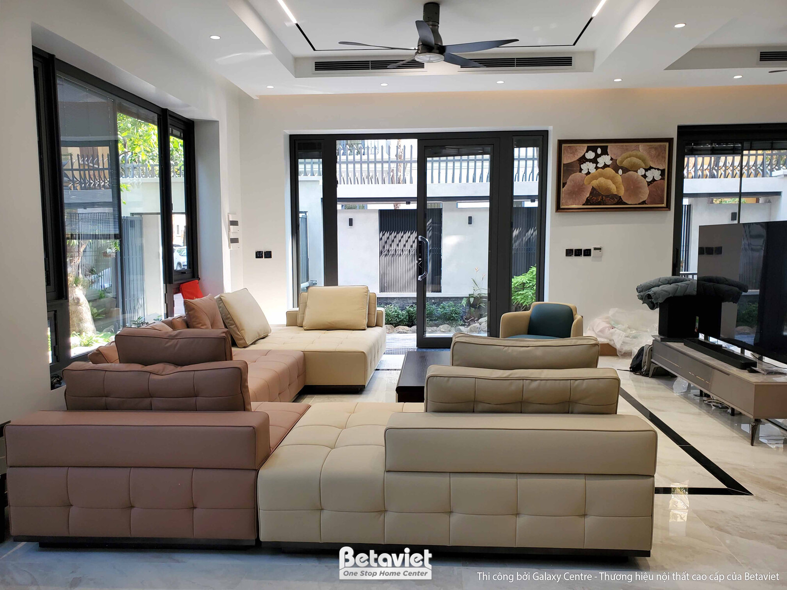 Phòng khách rộng rãi nhìn từ góc, bố trí bộ sofa lớn màu be và nâu nhạt. Trần thạch cao giật cấp với đèn âm trần, sàn lát…