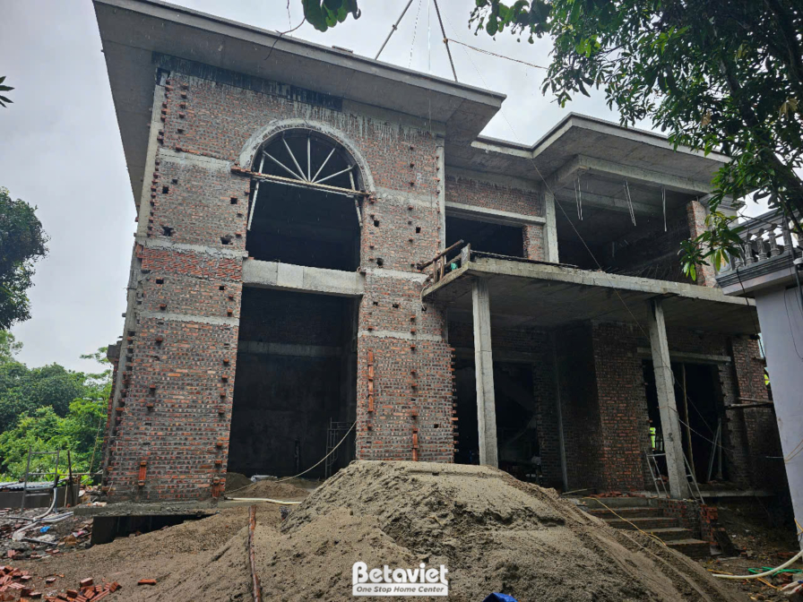 Mặt tiền chính của biệt thự đang trong giai đoạn xây tường gạch và định hình kết cấu. Các ô cửa sổ và cửa chính hình vòm l…