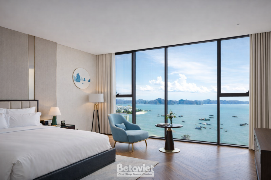 Phòng ngủ view vịnh biển đảo trực diện, giường ga trắng gối xanh navy, ghế bành xanh dương đặt cạnh cửa sổ, bàn tròn nhỏ…