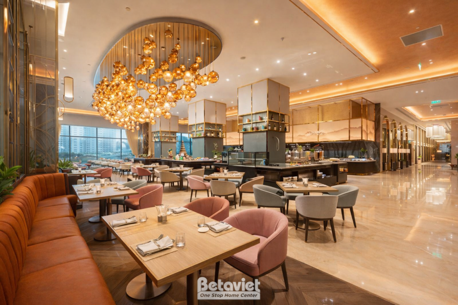 Nhà hàng buffet với cụm đèn chùm bóng tròn treo thành khối màu vàng đồng, bàn ghế bọc nệm cam đất và hồng phấn, sàn đá gra…