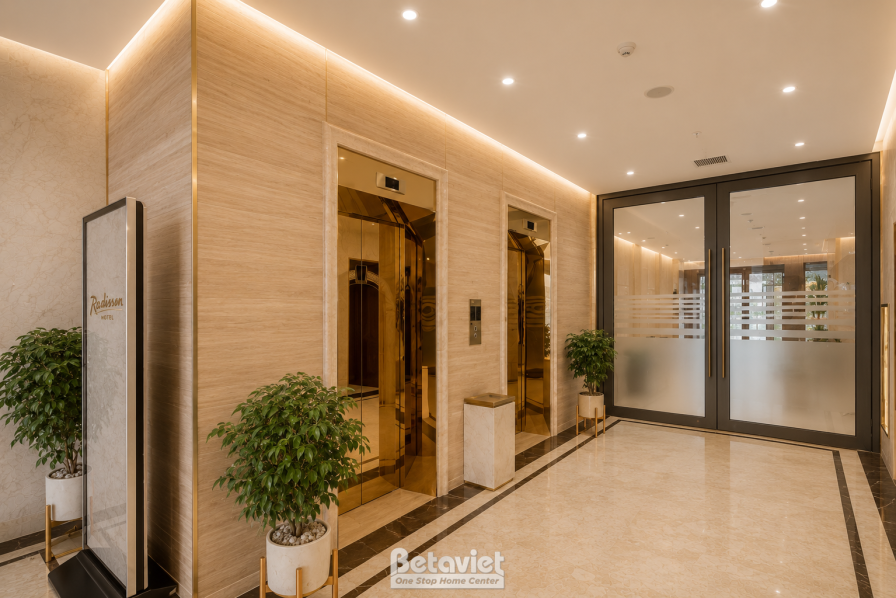 Hành lang khu thang máy ốp gỗ tông nâu sáng, cửa thang máy inox sáng bóng đặt song song, sàn đá marble kẻ line viền tối, c…
