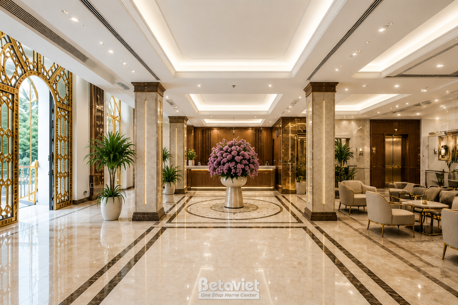 Sảnh lễ tân nhìn từ cửa vào, nền đá marble vân kem lát họa tiết viền hình học, hàng cột ốp vàng đồng chạy dài hai bên. Cổn…