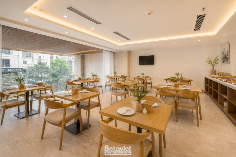 Khu vực bàn ăn sát vách kính lớn đón ánh sáng tự nhiên và view phố bên ngoài. Bàn ghế gỗ tự nhiên tông vàng nhạt bố trí th…