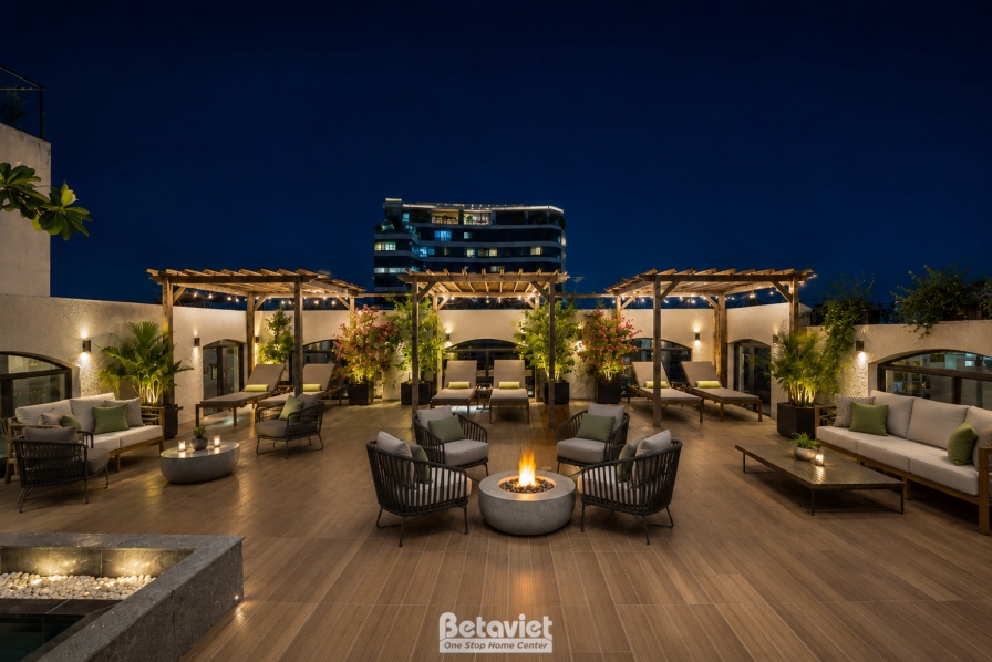 Góc khác của khu lounge sân thượng ban đêm, bàn lửa tròn đặt giữa cụm ghế đan dây đệm xanh rêu, hàng chậu hoa giấy đỏ điểm…
