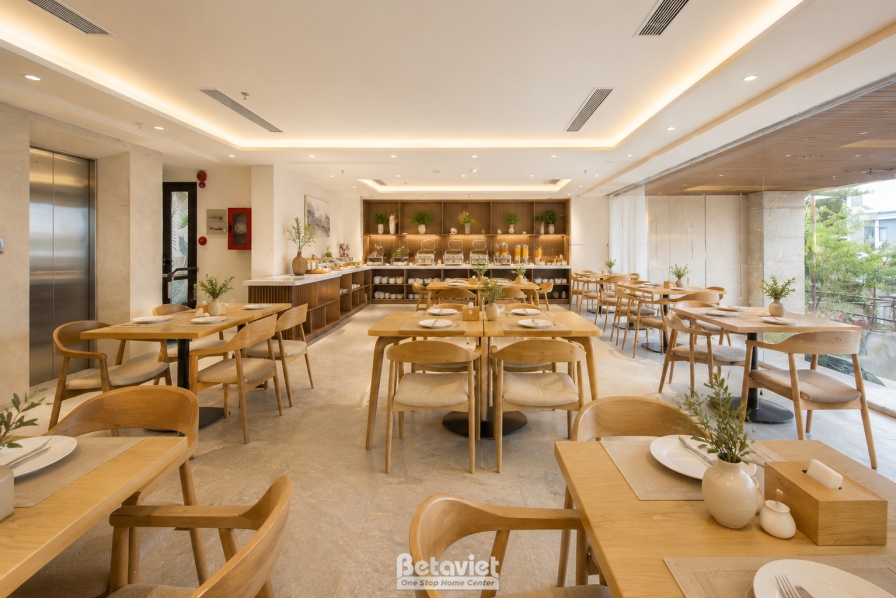 Toàn cảnh nhà hàng rộng với nhiều dãy bàn ghế gỗ, quầy buffet dài phía cuối, khu vực sảnh thang máy liền kề. Ánh sáng tự n…
